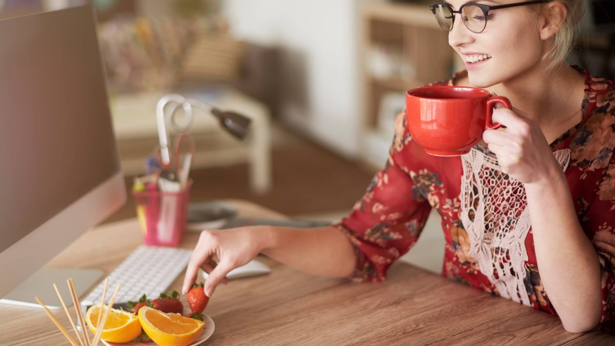 Mulher jovem, loira e com óculos de grau tomando um chá e comendo frutas sentada em frente de mesa