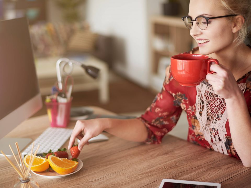 Mulher jovem, loira e com óculos de grau tomando um chá e comendo frutas sentada em frente de mesa