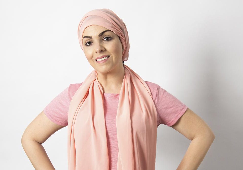 Mulher jovem, branca, com uma blusa branca de manga comprida e lenço rosa na cabeça, sorrindo
