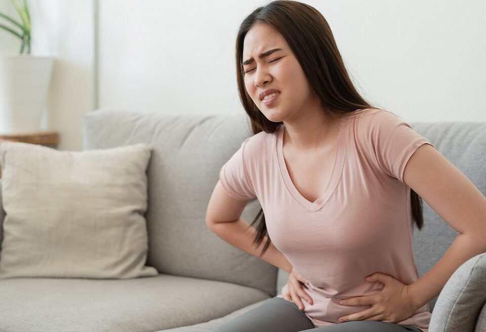 Mulher, jovem, oriental, sentada em sofá, com mãos na região do ventre por causa de dor devido endometriose