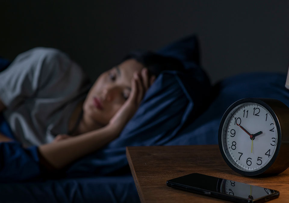 Mulher asiática jovem deitada em cama, com dificuldade para dormir, e despertador ao lado