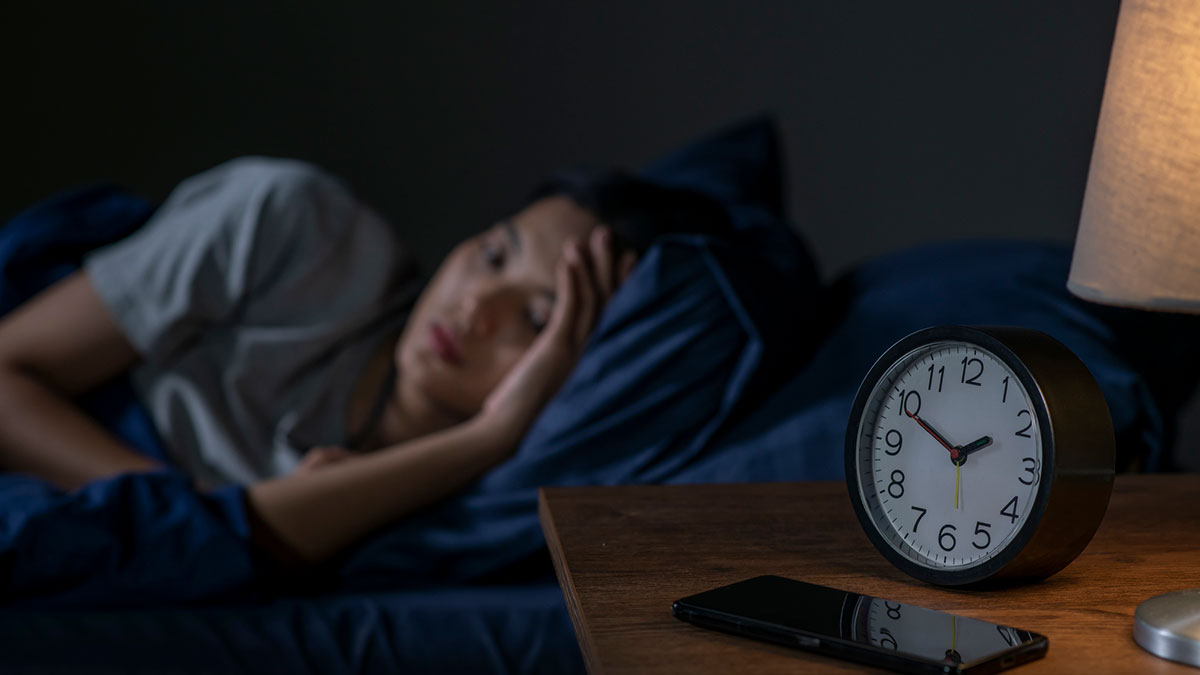 Mulher asiática jovem deitada em cama, com dificuldade para dormir, e despertador ao lado